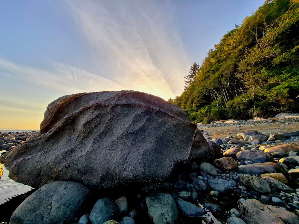 A large rocks sits at the oceans edge, worn by many tides past. Ripples wind down the rocks face as the sun sets in the distance behind it. 