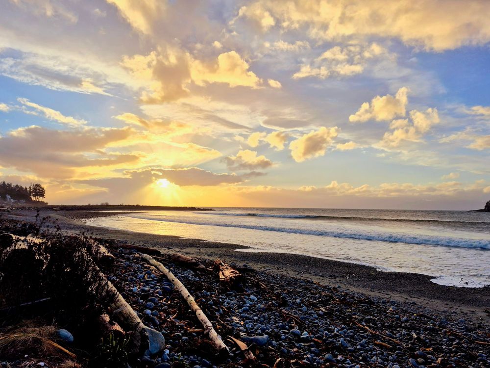 Sunrise on the beach, the sun bursts through the clouds with glorious sun beams. The calm ocean waves roll onto shore, the blue sky above is scattered with white puffy clouds.