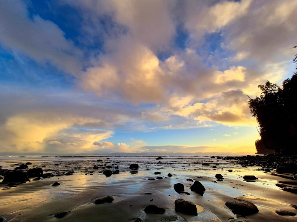 The beach is illuminated with beautiful light, rocks litter the sandy shore as the clouds dance above. 