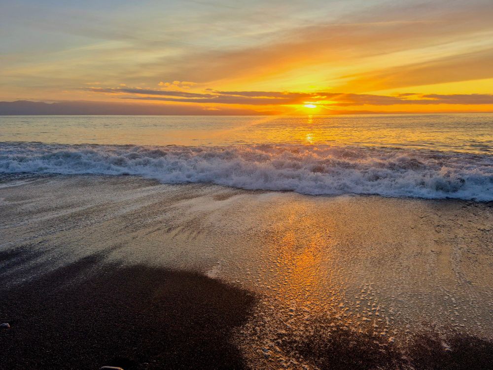 Sunset over the Pacific ocean, the sun just resting above the horizon. A big wave crashes to shore.