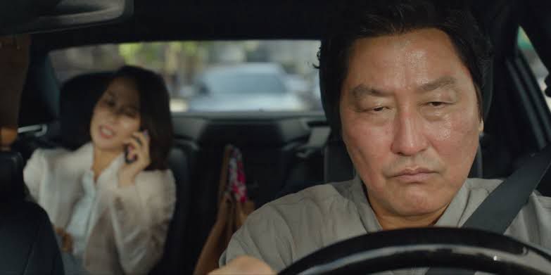 A cena do filme Parasita (2019), dirigido por Bong Joon-ho, mostra o personagem Kim Ki-taek (interpretado por Song Kang-ho) dirigindo enquanto a Sra. Park (Cho Yeo-jeong), elegante e distraída, fala ao telefone no banco de trás. A imagem simboliza a relação de servidão entre as classes retratadas no filme: Ki-taek, o pai da família pobre, trabalha como motorista particular para a família rica dos Park. Sua expressão séria e concentrada contrasta com a postura relaxada da passageira, sugerindo a tensão latente entre patrão e empregado. A cena exemplifica como o filme critica as desigualdades sociais e o abismo entre ricos e pobres na Coreia do Sul contemporânea.