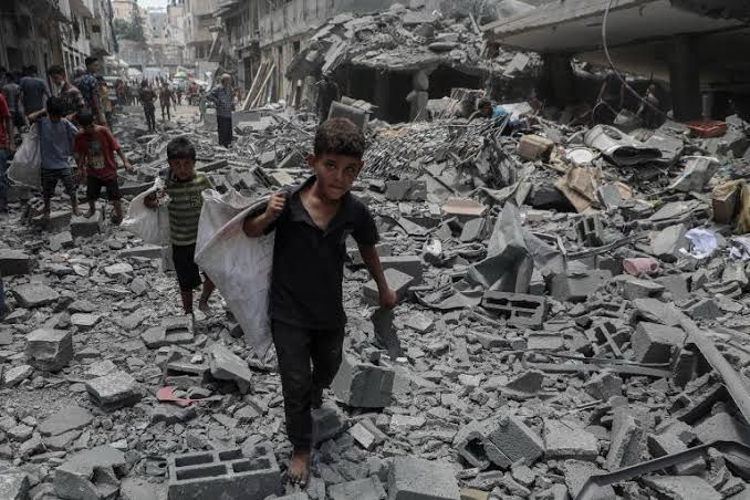 A imagem mostra uma cena devastadora em Gaza, com crianças caminhando descalças sobre escombros e destroços de concreto, carregando sacos. Ao fundo, edifícios estão destruídos ou severamente danificados, evidenciando o impacto de intensos bombardeios. Pedaços de tijolos, blocos e metal retorcido cobrem o chão, criando um cenário caótico. Outras pessoas aparecem ao longe, também atravessando a área destruída, possivelmente em busca de pertences ou ajuda. A atmosfera é de destruição generalizada e sofrimento humano, ressaltando as consequências humanitárias e sociais de conflitos armados prolongados.