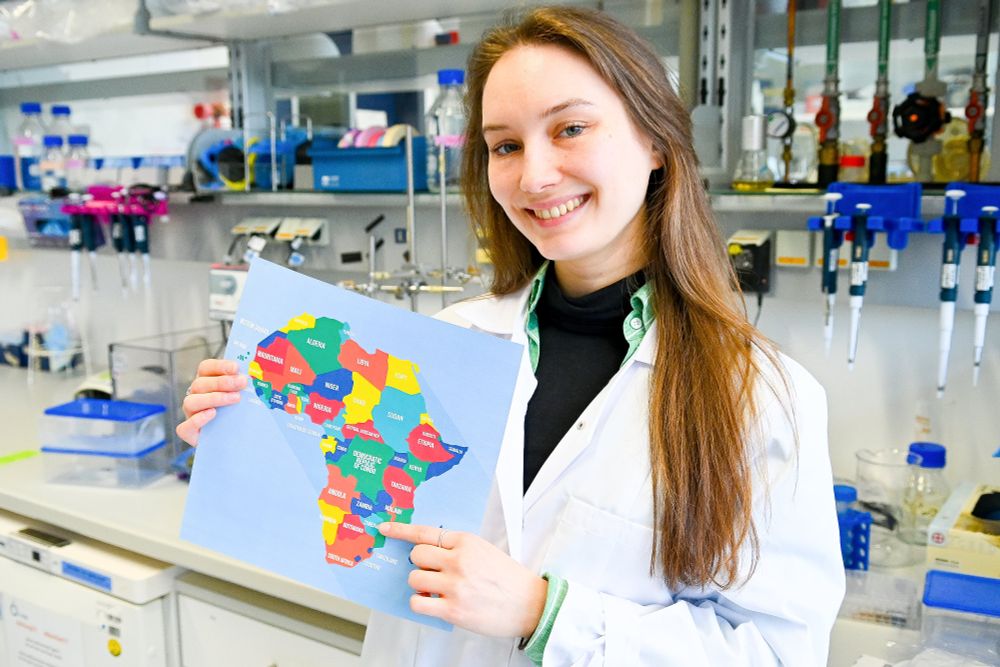 The photo shows a smiling Noemi Iwicki in a lab coat, with the laboratory in the background. She holds a colourful printout of the African continent and points to Zimbabwe.
