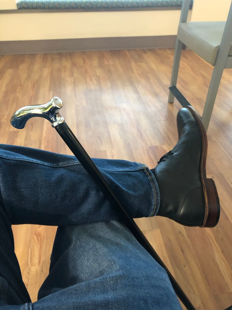 A man’s left leg up and crossed over resting his right knee, wearing black leather Italian dress half boots, blue jeans, and with the finishing touch of one of his black wood walking canes with a silver derby handle resting up against the right side of his right leg, right back against the back side of his raised and crossed over left leg just behind his right knee, keeping the cane from falling. Functional style, or something like it, for a younger guy who’s been forced by his medical team to walk with a cane. So, make the most of it, lol.

