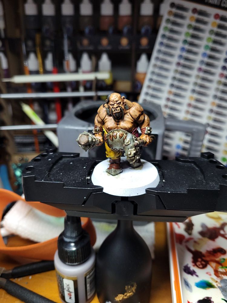 What you see is a Warhammer ogre miniature on a painting stand. It has olive skin, and black hair with a terrible mustache and a shaved head with a single ponytail. Otherwise this fat human like creature hides a lot of muscle under minimal clothing, including light wrist armor, an iron and brass plate over its gut, yellow and burgundy pants, and leather boots that have steel plate toes. This ogre holds two Stone and metal clubs with wood handles, and wears two stone slab earrings.