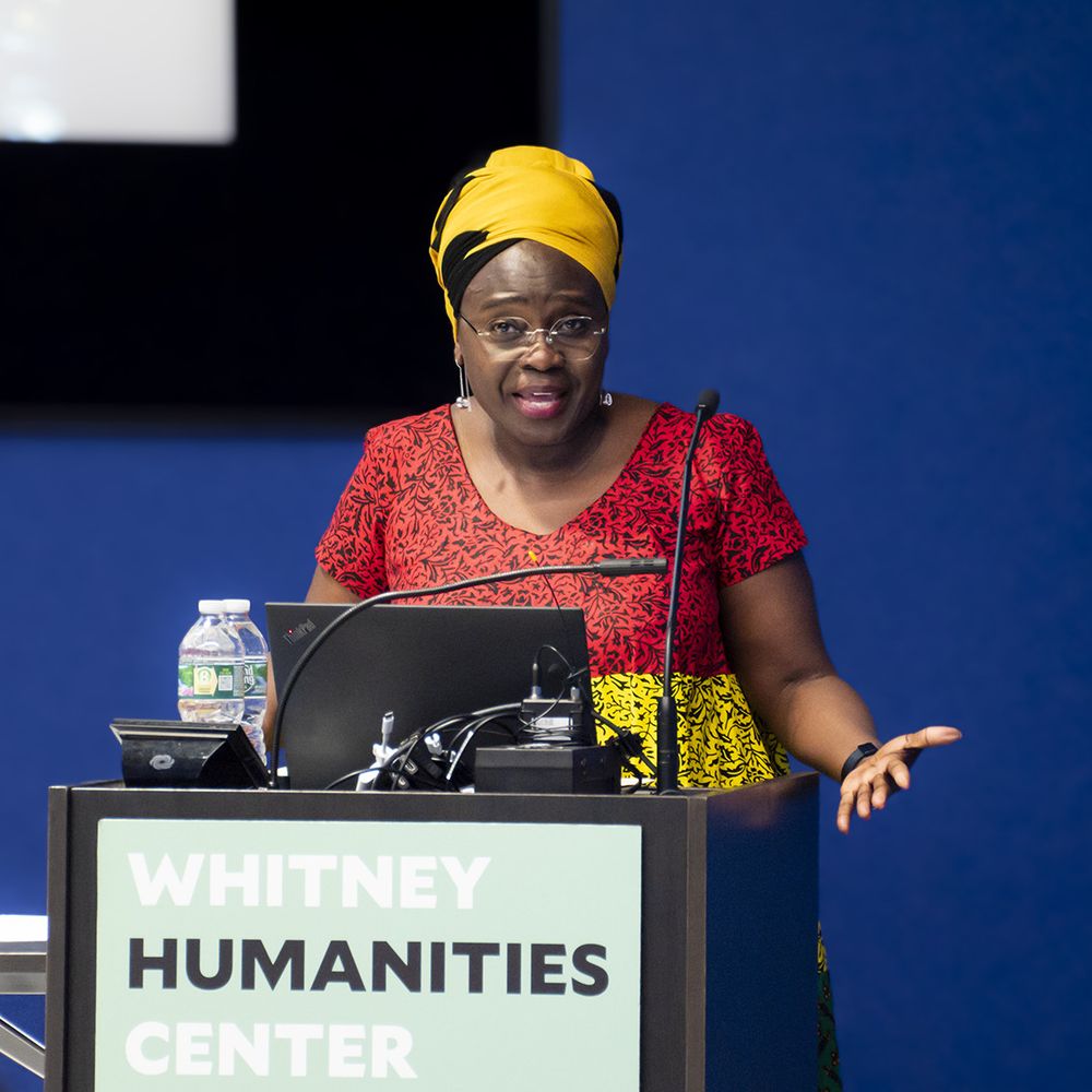 A woman wearing a yellow African headwrap and red and yellow print dress speaking to an audience from behind a lectern.