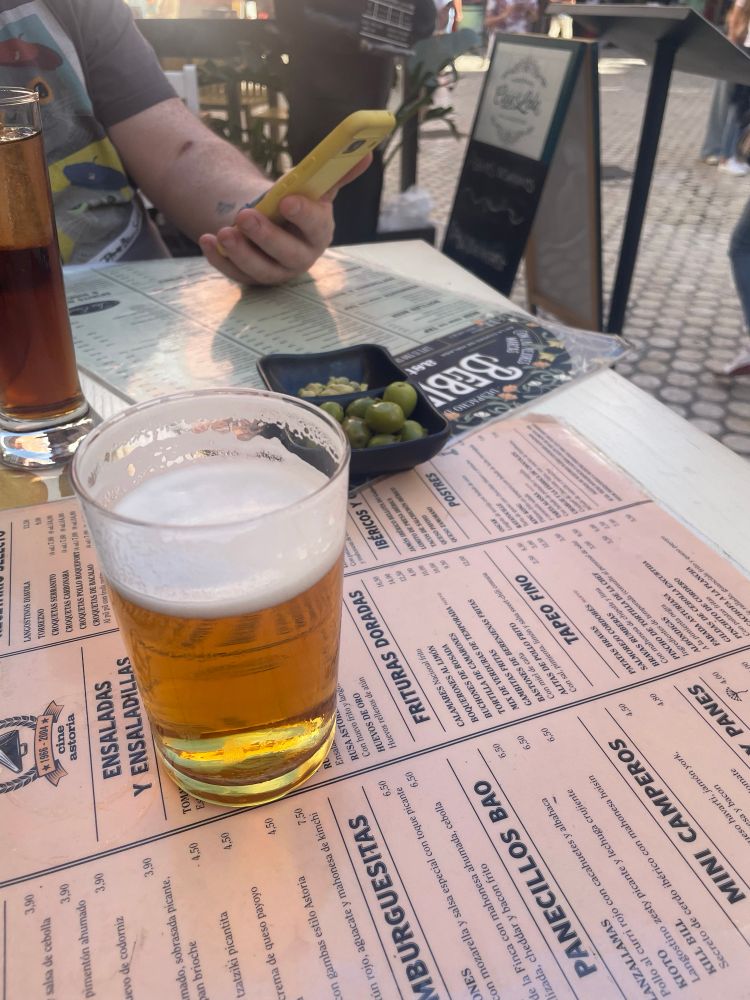 A small beer (cana) and a bowl of olives sitting on a laminated menu in Spanish, on an outdoor table by a taberna. It’s very sunny. 