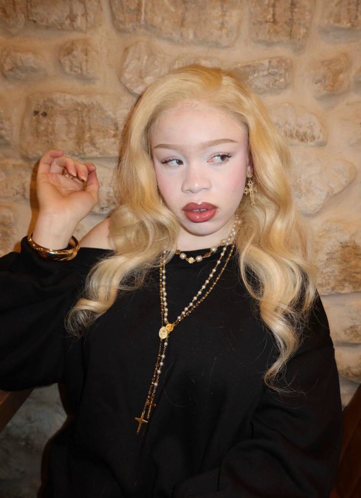 Photo of a black woman with albinism, blonde hair and fair akin wearing a black shirt and in front if a stone wall