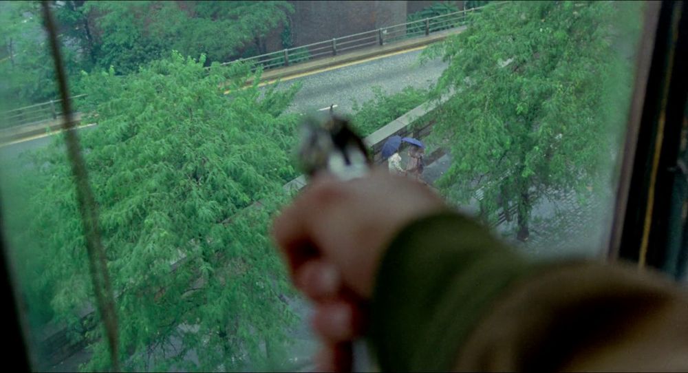 TAXI DRIVER: Travis' out of focus hand holding a gun, aiming it through a window in a high hotel floor, the trees bright green, and two women talking on the sidewalk