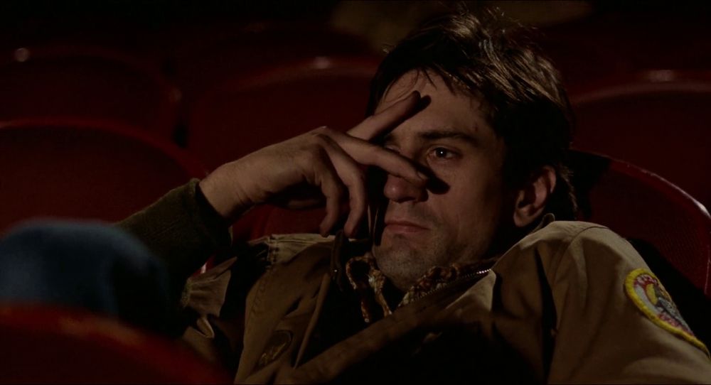 TAXI DRIVER: Travis sitting in a movie theater, slouched low in the seat, holding up his hand to his face, making a V sign over his eyes