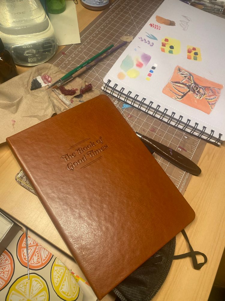 A picture with a leather bound notebook that states, “The Book of Good Times” on the cover. The notebook lays atop a kn95 mask and next to a watercolor painting. 
