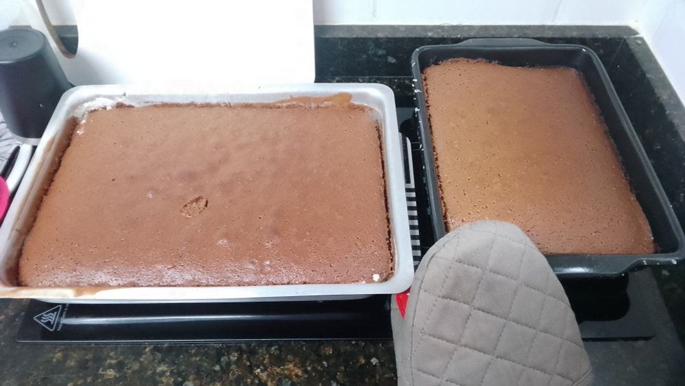 Alt text:  
Duas formas retangulares de bolo de chocolate recém-assado estão sobre uma bancada de cozinha com tampo de granito preto. O bolo da esquerda tem uma pequena marca afundada no centro, possivelmente feita para verificar o ponto de cozimento. Ambos os bolos têm superfície lisa e brilhante, indicando que foram assados uniformemente. Uma luva térmica acolchoada aparece em primeiro plano, sugerindo que os bolos foram retirados do forno há pouco. Ao fundo, há azulejos brancos na parede, compondo um ambiente de cozinha moderna.