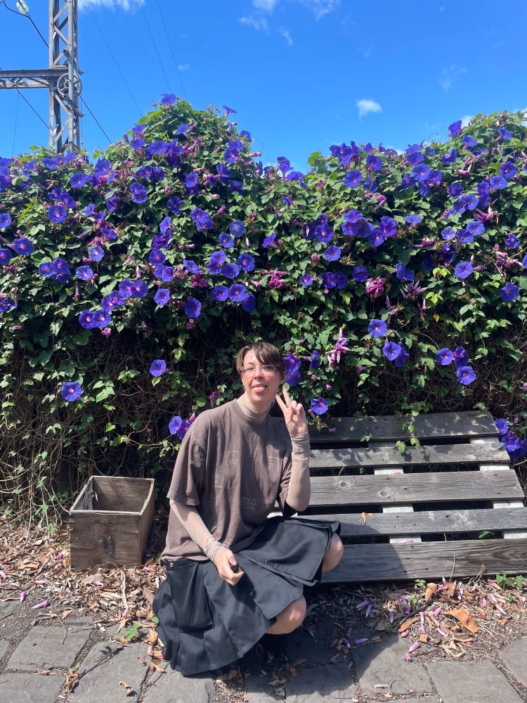 Talzir (user) poses crouched in front of butterfly pea hedge and wooden palette on cobblestones. They wear an oversized brown T-shirt and pleated black skirt. They throw a peace sign, and look silly in their little outfit. 