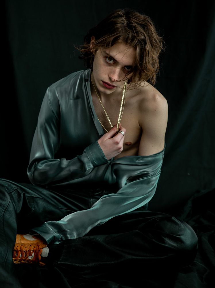 White male model with slightly long light brown hair. Wearing a blue silk shirt off of one shoulder and leather pants against a black fabric backdrop.