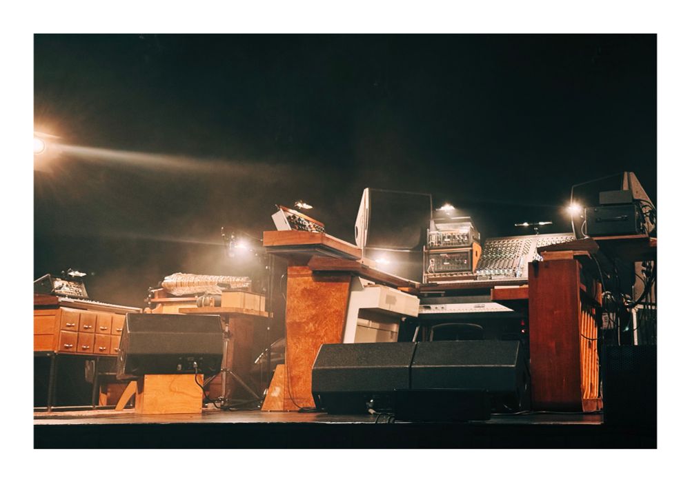 A dimly lit stage setup featuring a variety of analog synthesizers, wooden desks, a mixing console, and rack-mounted audio equipment. Warm light beams cut through the haze, highlighting the retro-futuristic aesthetic of the instruments and furniture. Large speakers sit at the front edge of the stage.