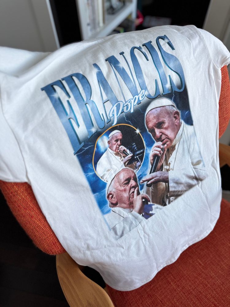White T-shirt draped over a red chair, featuring a dramatic graphic of Pope Francis in multiple poses with the text “FRANCIS” and “Pope” in stylized blue lettering, set against a lightning-filled background.