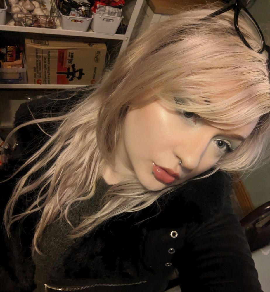 Selfie of a person with long blonde hair, glasses in their hair, looking up at a camera with lots of ingredients like ramen and garlic on a shelf behind