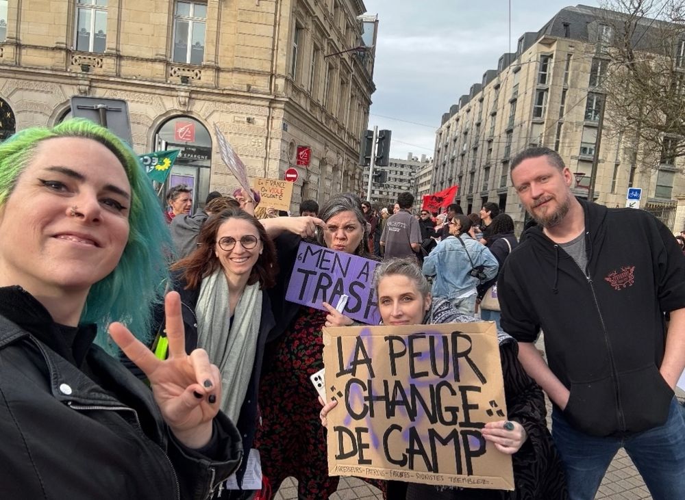 Photo de manifestation pour la journée de lutte des droits des femmes et minorités.

Au premier plan mon amie Rebecca, connue sous le pseudo de Valmyr Salt (tatoueuse), c'est elle qui prend la photo en selfie, et fait un V avec les doigts de la main libre, à sa gauche notre Caroline, à sa gauche mon amie et collègue Bloody Mary (tatoueuse) qui tient une pancarte "Men are trash", à sa gauche moi, Mymy Magma (tatoueuse) qui tient une pancarte "la peur change de camp", à ma gauche mon conjoint Axel, qui rejoint régulièrement la lutte. En fond, d'autres manifestantes avec banderoles et pancartes.