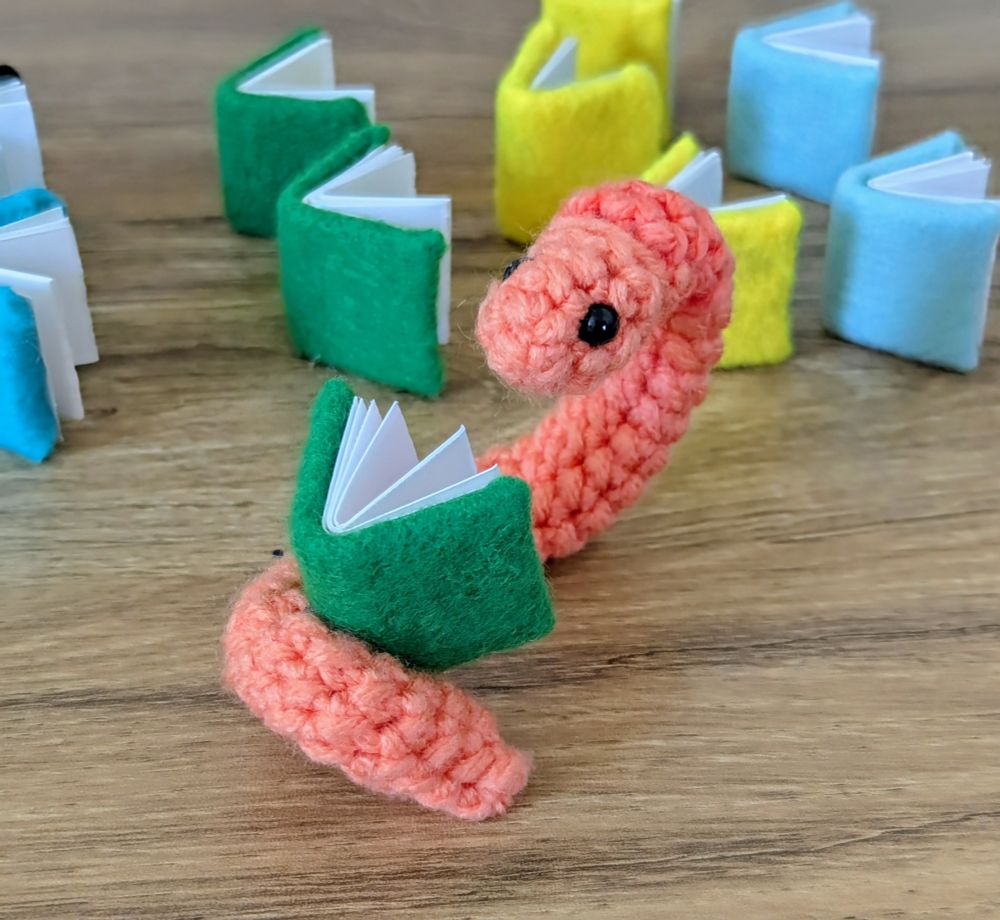 A crocheted worm reading a tiny book made of felt and paper. It is surrounded by similar tiny books on a wooden table.