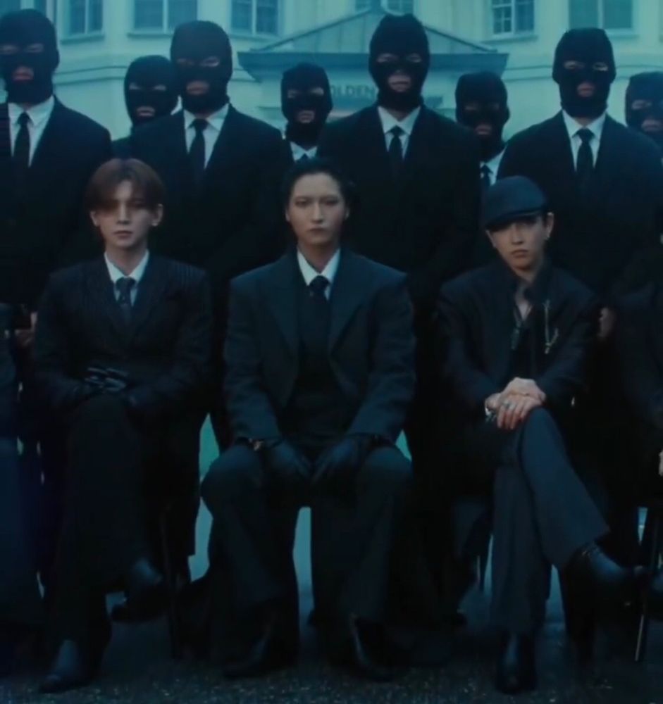 A screen shot from the Ice in my teeth MV trailer of Seonghwa with his hair slicked back, sitting, looking serious next to Yeosang and hongjoong, outside a mansion. There’s a lot of men wearing black masks and suits standing behind them. They look like mob bosses tbh. 