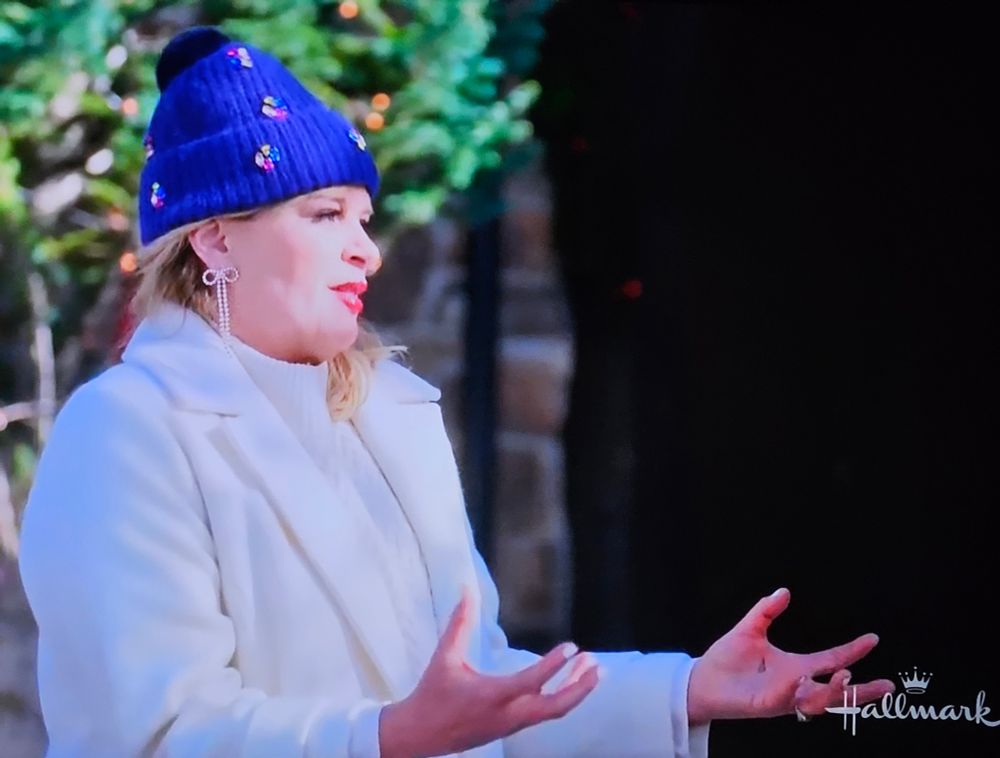 A woman standing outside in a white coat and blue hat. She has dangling diamond earrings on that are supposed to be a Christmas bow I believe but at first glance they looked like a dick and balls to me.