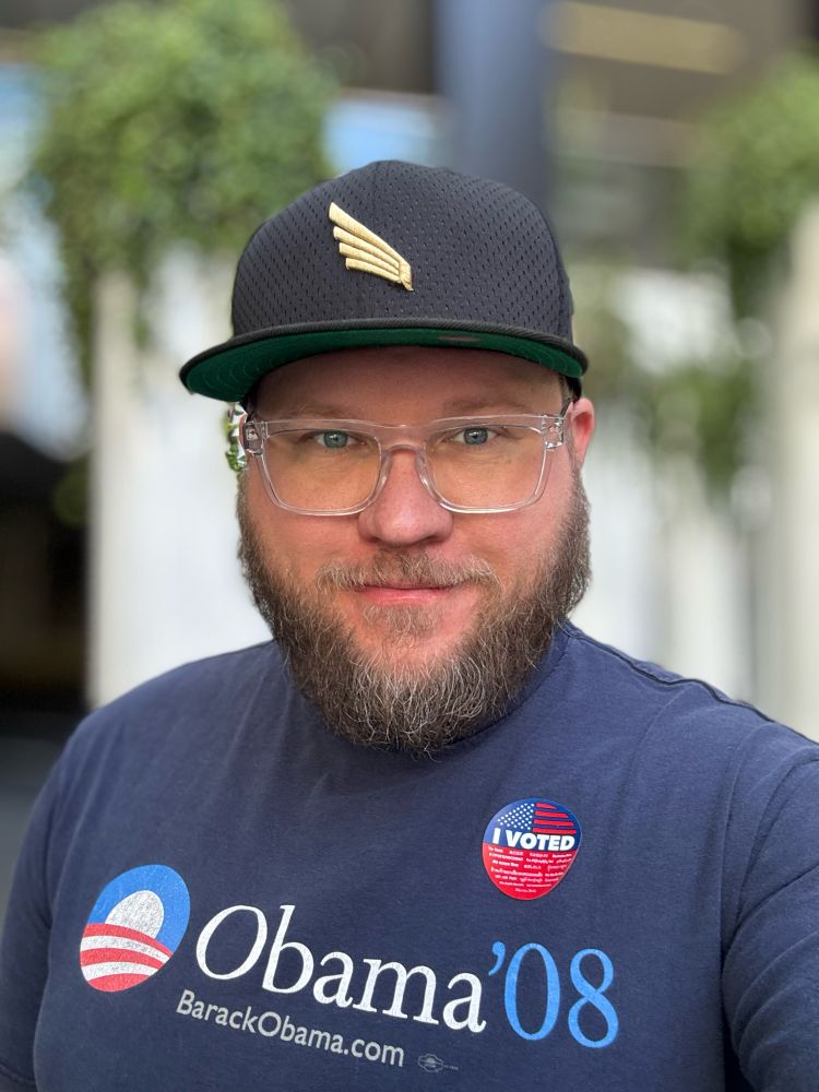 Selfie of me wearing the same blue Obama ‘08 T-shirt with an I voted sticker and an LAFC baseball cap. I am 39 and have a beard and clear framed glasses