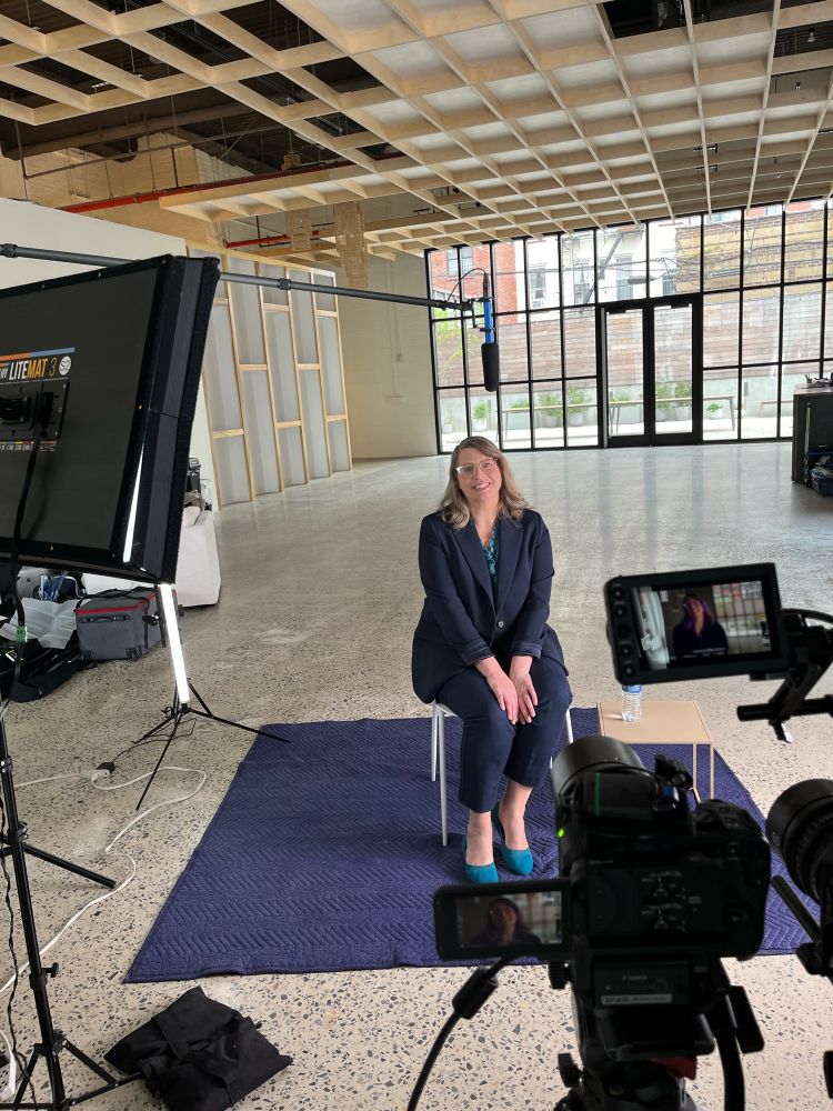 Woman in a navy blazer sits in a chair on a rug in the middle of a large airy room with cameras and equipment in the foreground 