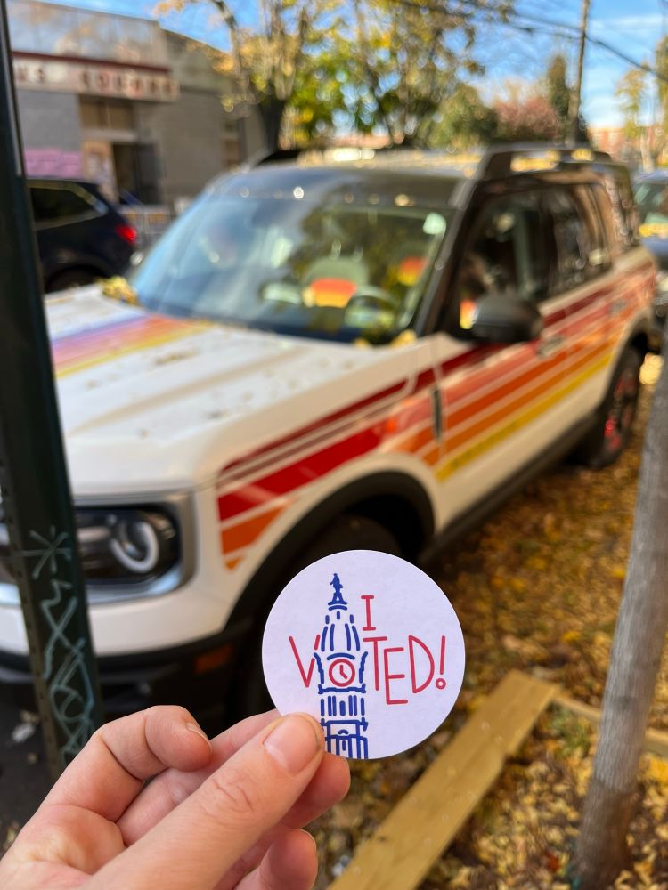 I voted! Sticker with city hall graphic; in the background is a white ford bronco with cool red orange and yellow stripes