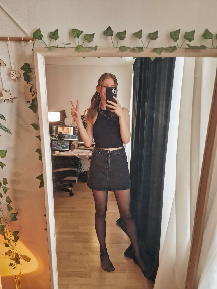dark blonde woman standing in front of the mirror taking a selfie, wearing a black skirt and black tank top.