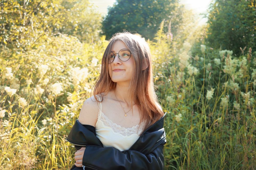 A portrait of Joy in front of a lush garden or meadow, she's looking up smiling and the sun is hitting her head. She's a brunette wearing a light tank top with lace and a black jacket.