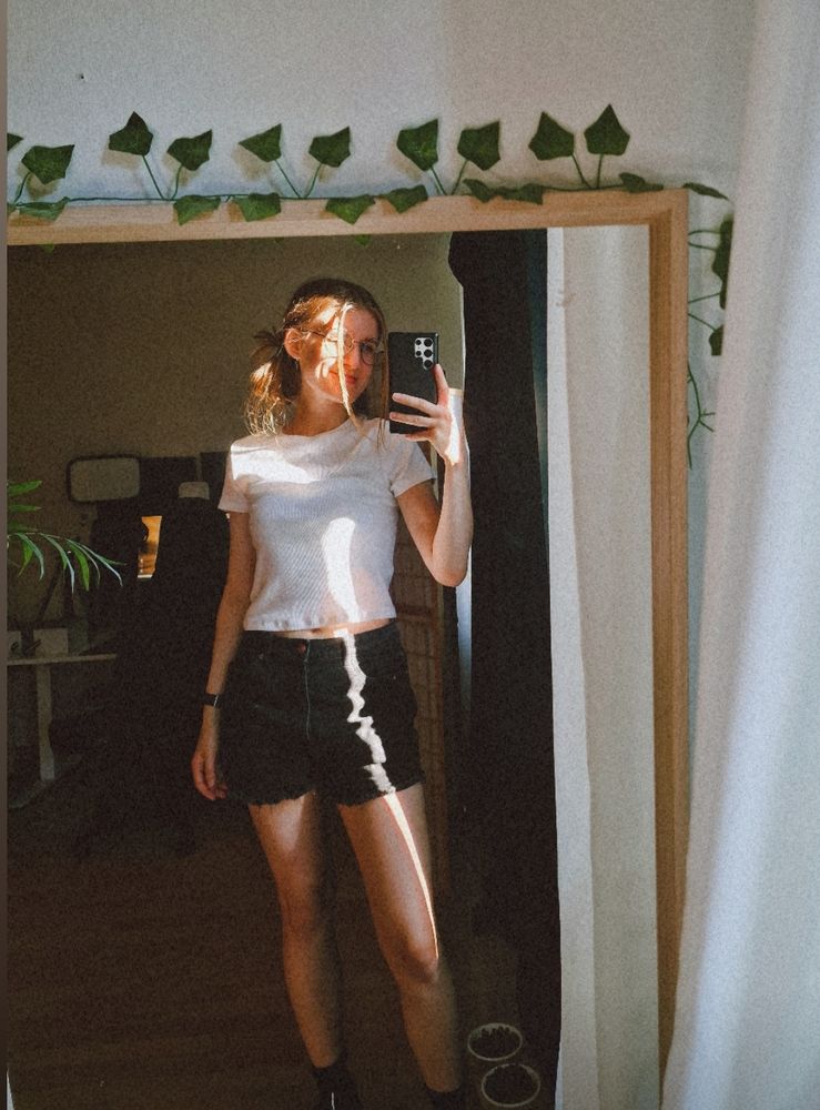 selfie of a woman with a white t-shirt and black shorts in front of a mirror. There's a sunlight beam across her body.