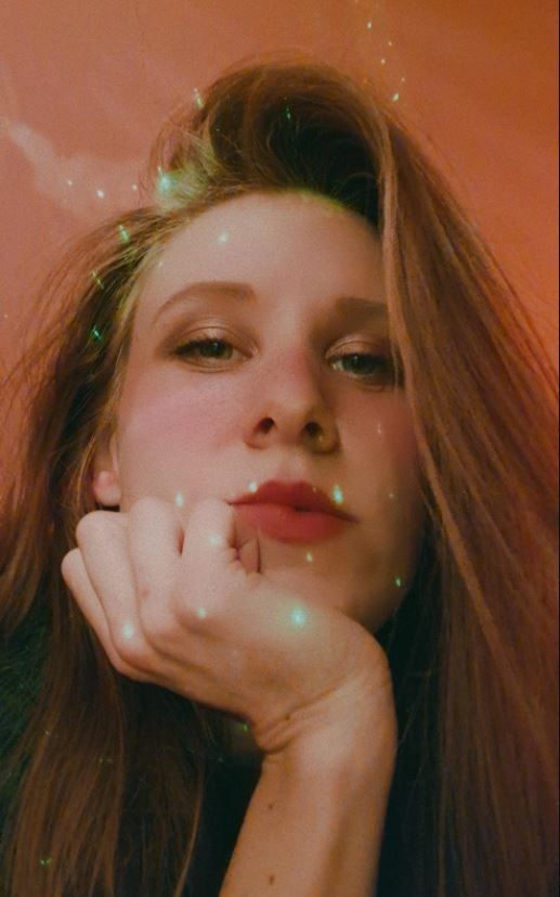picture of Joy, a women with light brown hair, looking into the camera with red lipstick and sparkly green light around her.