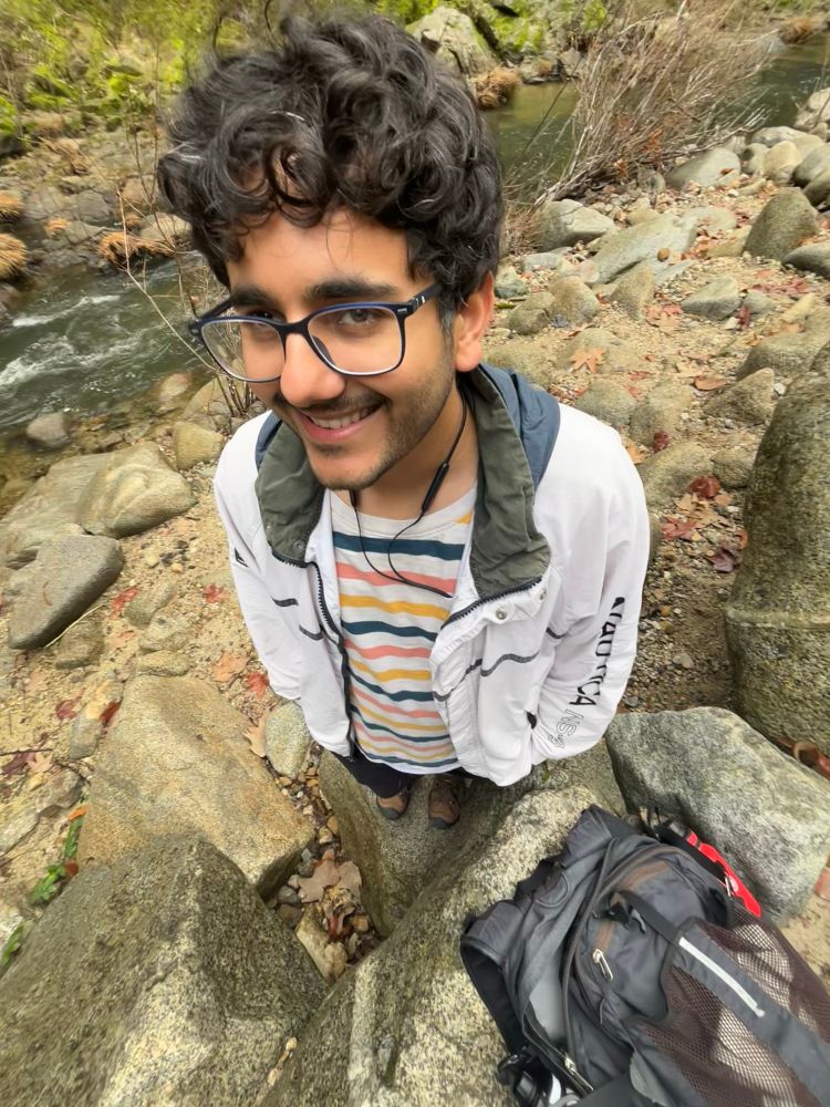 me at a stream. backpack on the ground, red and white water bottles on each side. wearing a white nautical with a line across the chest, a dark hood. horizontal lines tee and hiking boots.