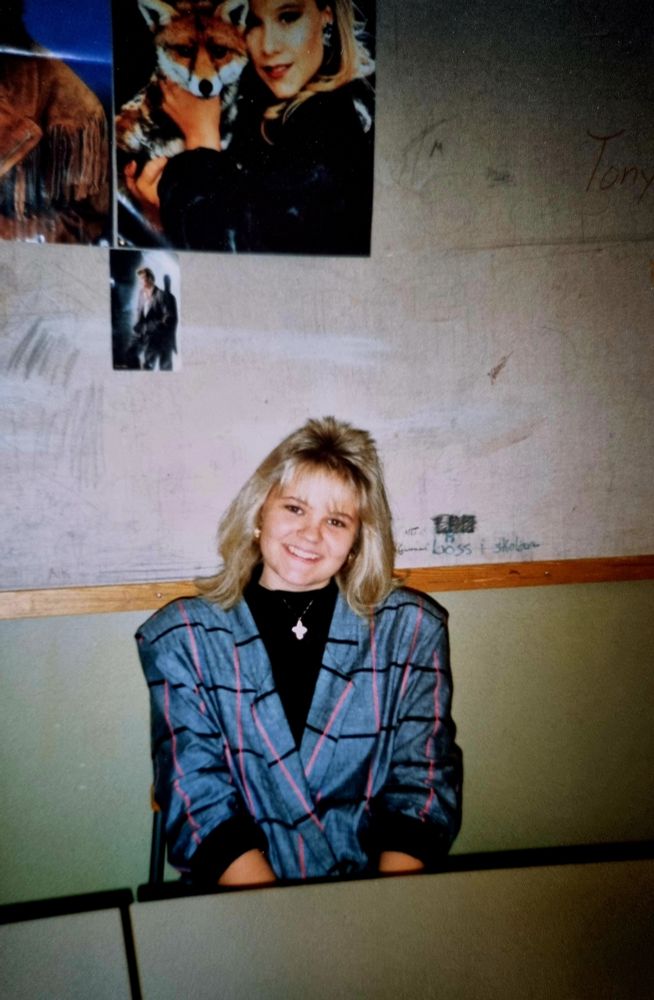 Blond tonårstjej med storutig kavaj sittandes vid en skolbänk med en affisch på vägen bakom med Samantha Fox som håller i en räv. 