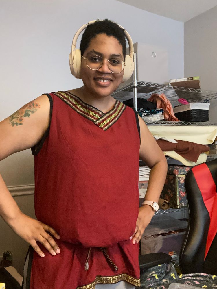 Me in a red medieval style tunic in red with golden and red trim around the collar and at the bottom. There’s a brown rope tied at my hips. I have my hands on my hips and I’m smiling at the camera. 