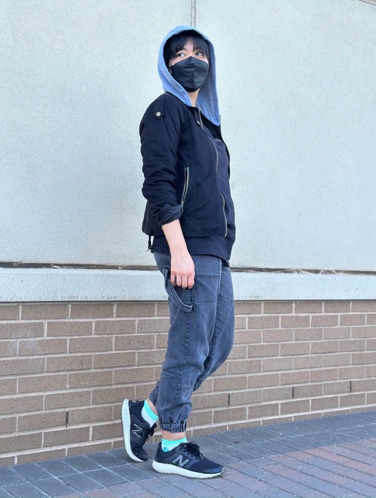 photo of a Mitsuki Koga cosplayer standing in front of a wall. She wears a black jacket with a gray hood, a black mask, baggy dark gray joggers, black sneakers, and green socks. He seems to be walking forward, looking over his shoulder.