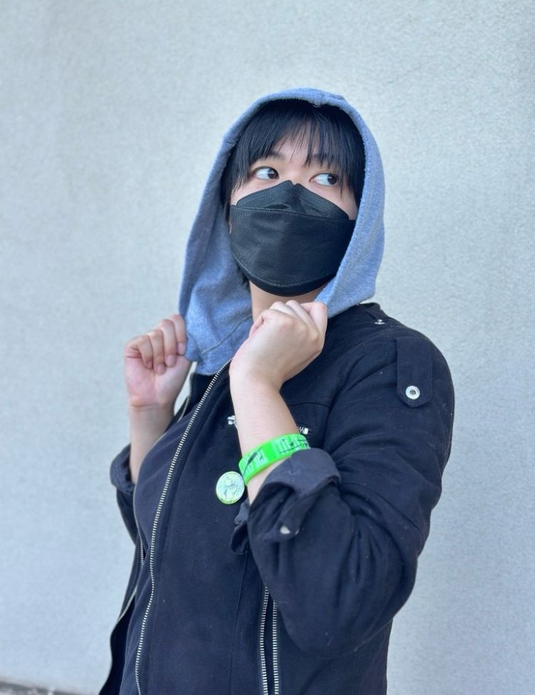 photo of a Mitsuki Koga cosplayer standing in front of a wall. He tugs the hood of his jacket forward. A green button is pinned to the front of her jacket, and she has a green wristband on her arm.