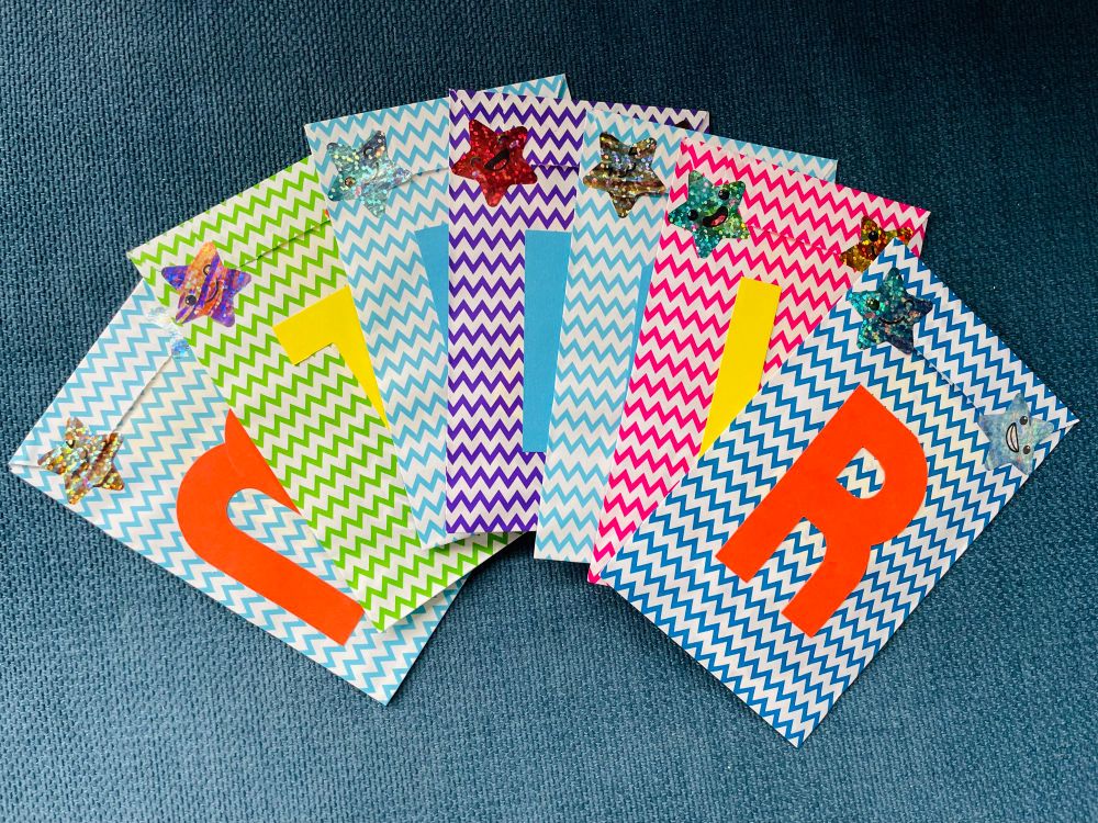 Photo of a set of paper bags in bright colours arranged on a blue background. Each paper bag has shiny star stickers on it and coloured initials.