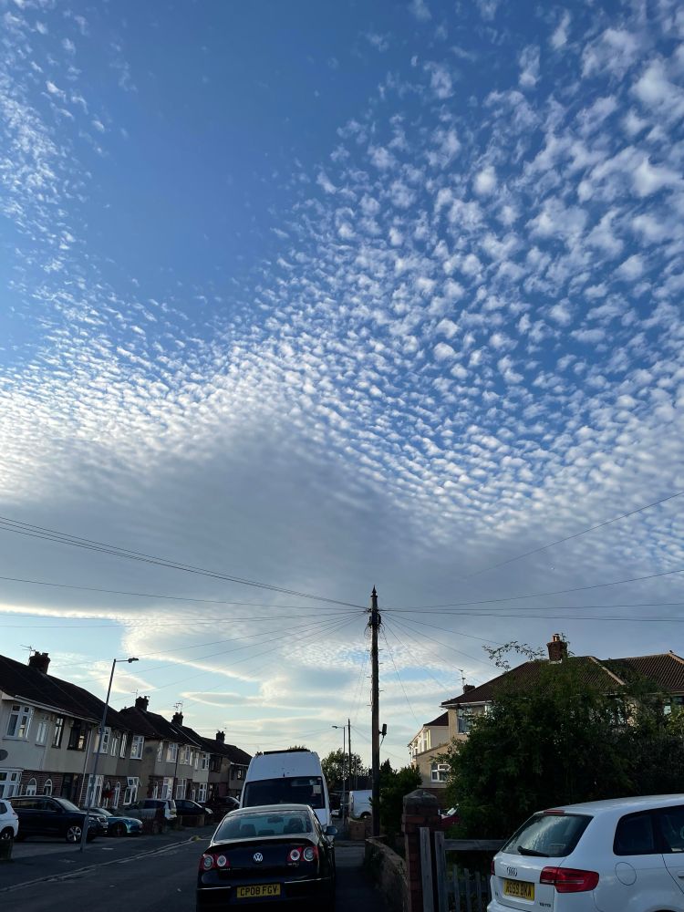 Photo of skies above a street in Bristol, there is a lot of pavement parking also in shot. The clouds are white and grey against a bright blue sky. They break up towards the top of the photo into increasingly small little clouds.