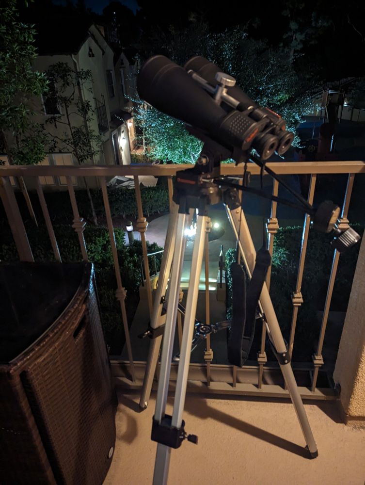 Celestron Skymaster binoculars on a metal tripod, set up on a small balcony.