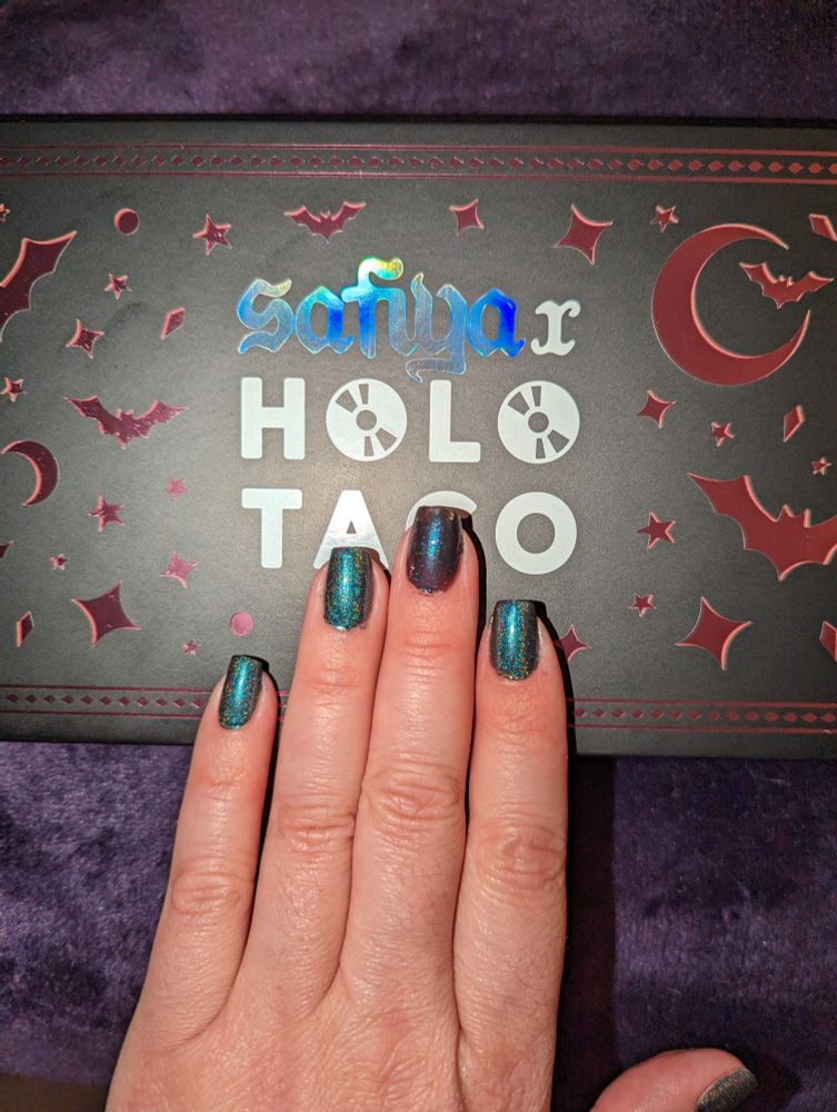 Swatching some of the new Holi Taco x Safiya collab nail polishes. 

A hand with short nails painted in two of the colors from the collection. Four of the nails on the hand are painted in Brucie, a muted dark teal with linear holo, the middle nail is painted in Bat B*tch, a black pearlescent with blue miro-shimmer. The hand is over the collection box which is black with reflective pinky-purple cut outs in the shape of bats and moons.