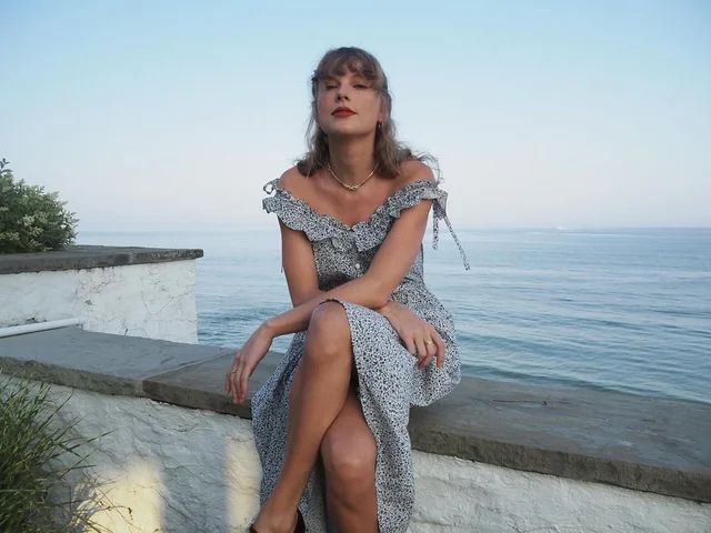 Taylor Swift from a past “belated Independence Day” post - she is sitting on a seat wall in front of a large body of water while wearing a ruffled off the shoulder patterned dress. 
