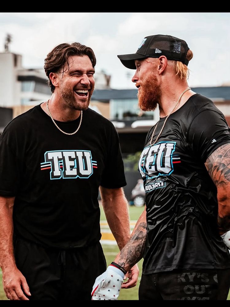 Travis Kelce is laughing with George Kittle. Both men are in black shorts and black TEU t-shirts, but Travis is dry while George is soaked
