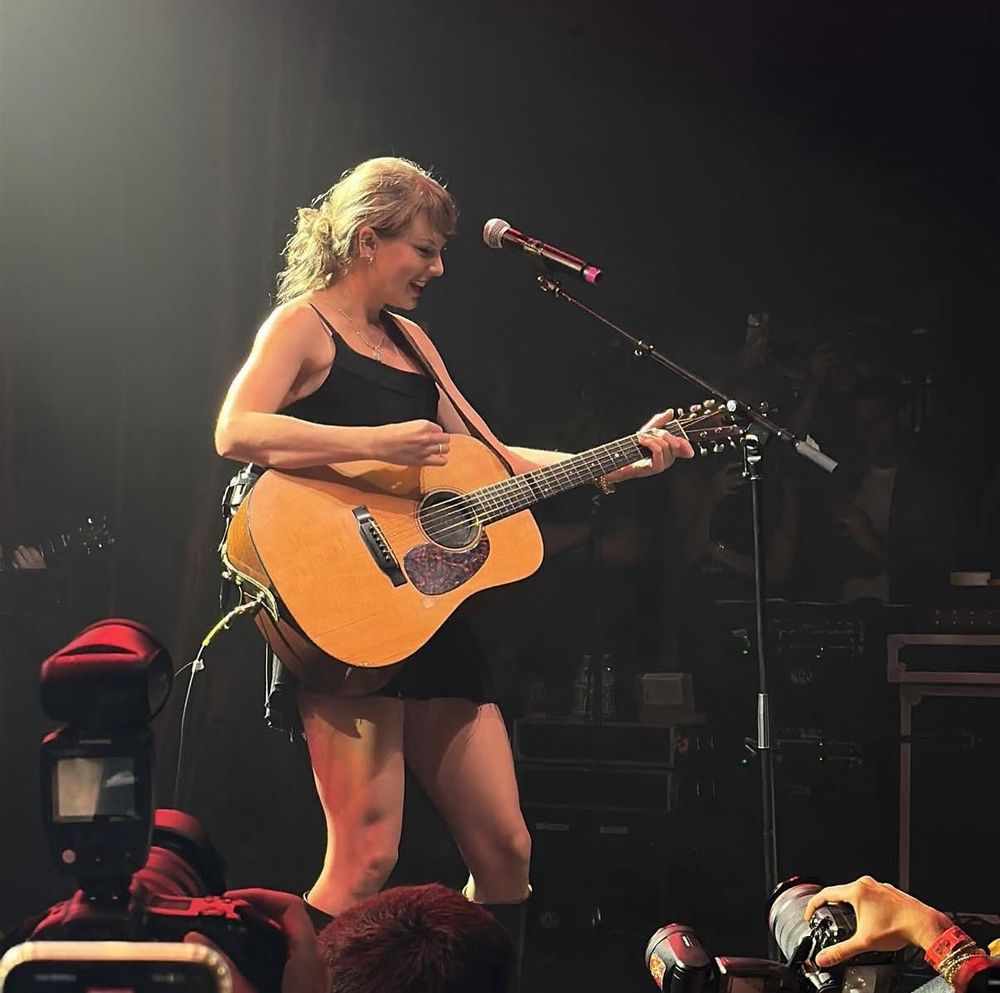 Taylor Swift on stage at Tight End University playing guitar and smiling