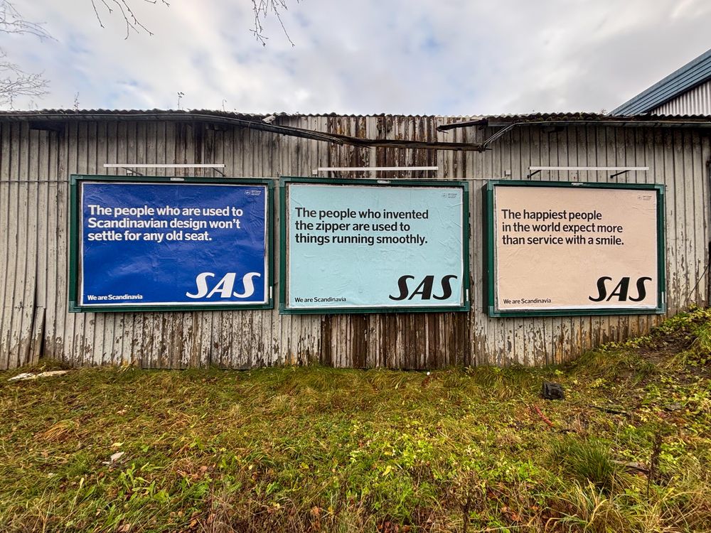 Tre engelskspråklige reklamer for SAS med tematikken "vi er fra skandinavia, vi er flinke" på en en nedslitt vegg hvor malingen flasser og takrenna er løs.