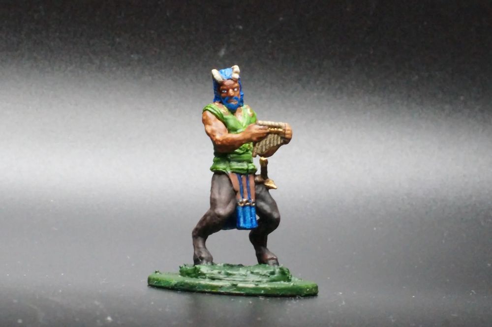 A satyr, a jolly/raucous fey creature in D&D. He has the upper body of a human and the lower body of a goat, but also has horns sprouting from his head. He has a green jerkin, blue hair and a blue beard, a blue and brown tabard, and carries a sheathed sword and a pan flute.