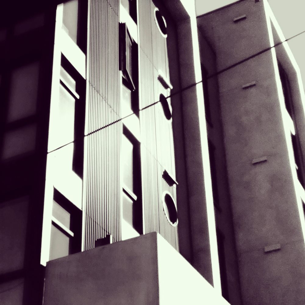 Magenta tinged image showing the exterior of a building shot at an angle emphasizing the variety of window shapes in late afternoon shadow.
