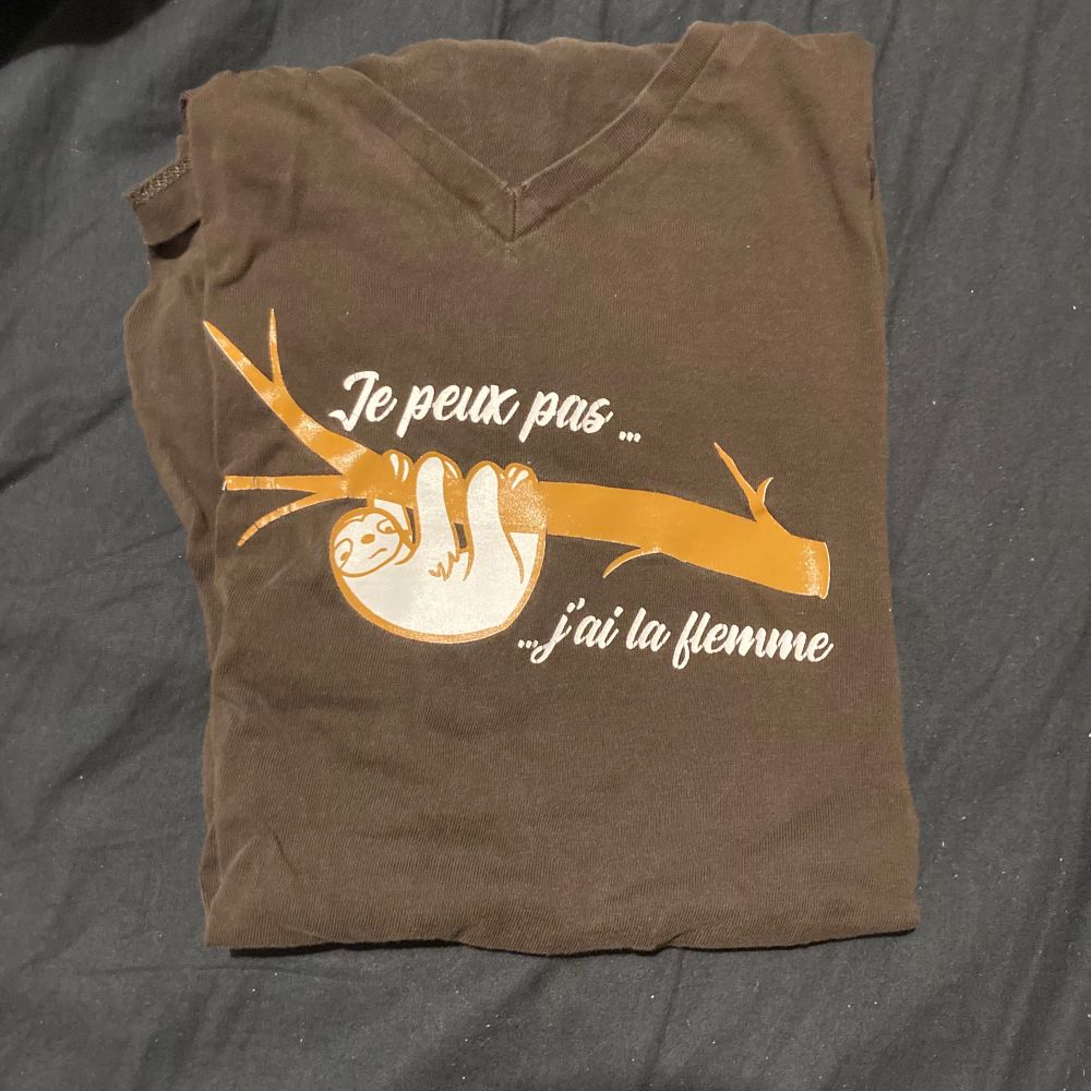 Le t-shirt marron plié sur lequel il y a un dessin de paresseux accroché sur une branche entouré de phrase « Je peux pas… j’ai la flemme »