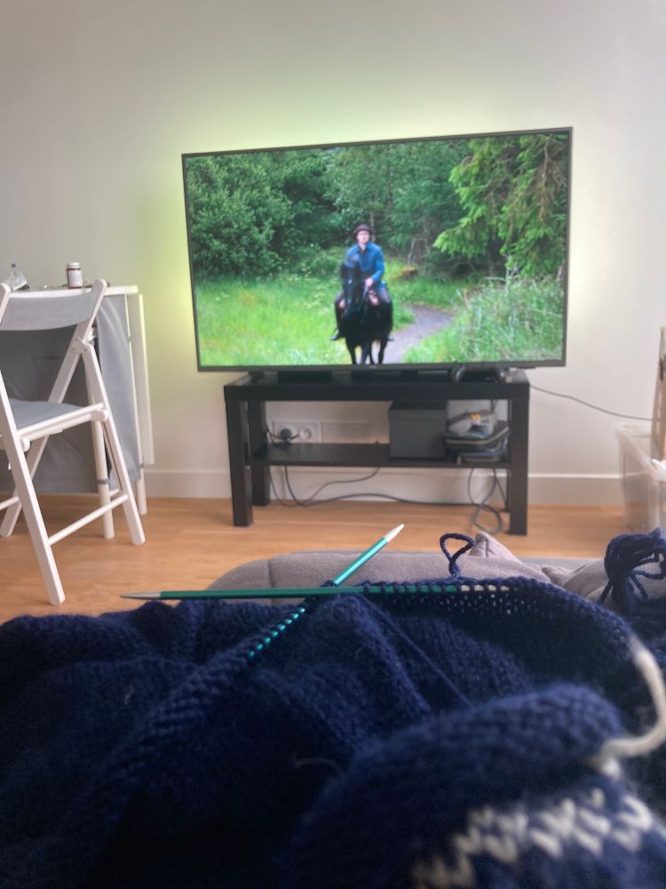 Tricot bleu foncé en cours en premier plan. La télé avec une scène de la série Outlander en arrière plan.