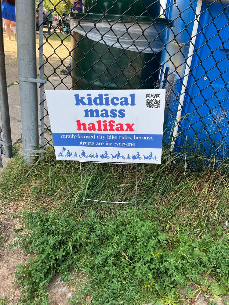 Sign for Kidical Mass Halifax stuck in a patch of grass which reads "kidical mass Halifax, Family-focused city bike rides, because streets are for everyone"